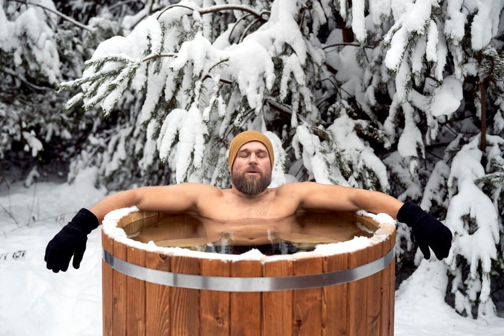 A Caucasian man sits in a wooden tub outdoors in a snowy forest.