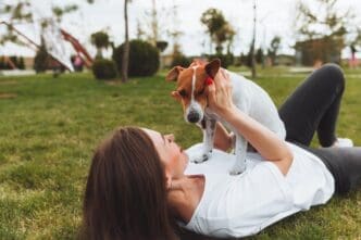A Caucasian woman lies on the grass in a park, playing with her Jack Russell Terrier.