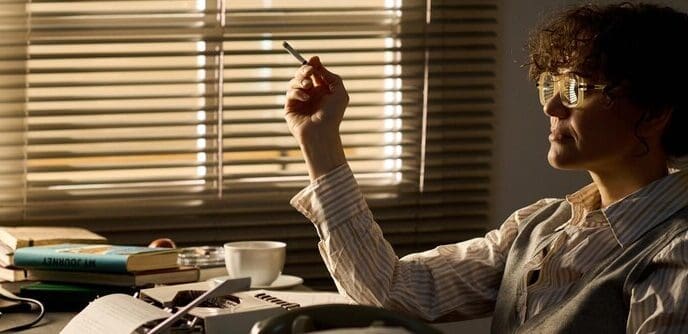 A young white woman sits at a desk, deep in thought, with a cigarette in her hand.