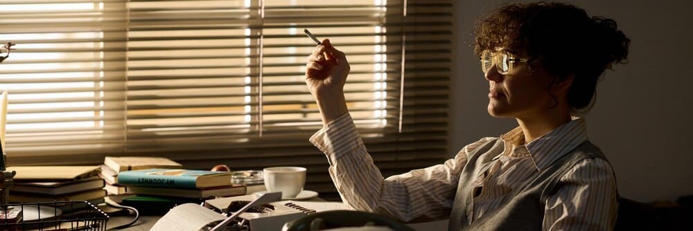 A young white woman sits at a desk, deep in thought, with a cigarette in her hand.