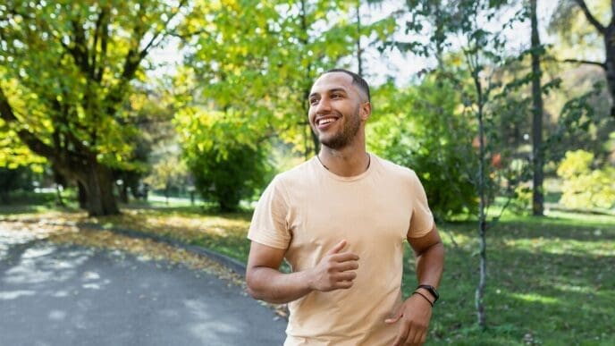 A smiling Hispanic man jogs through a sunny park.