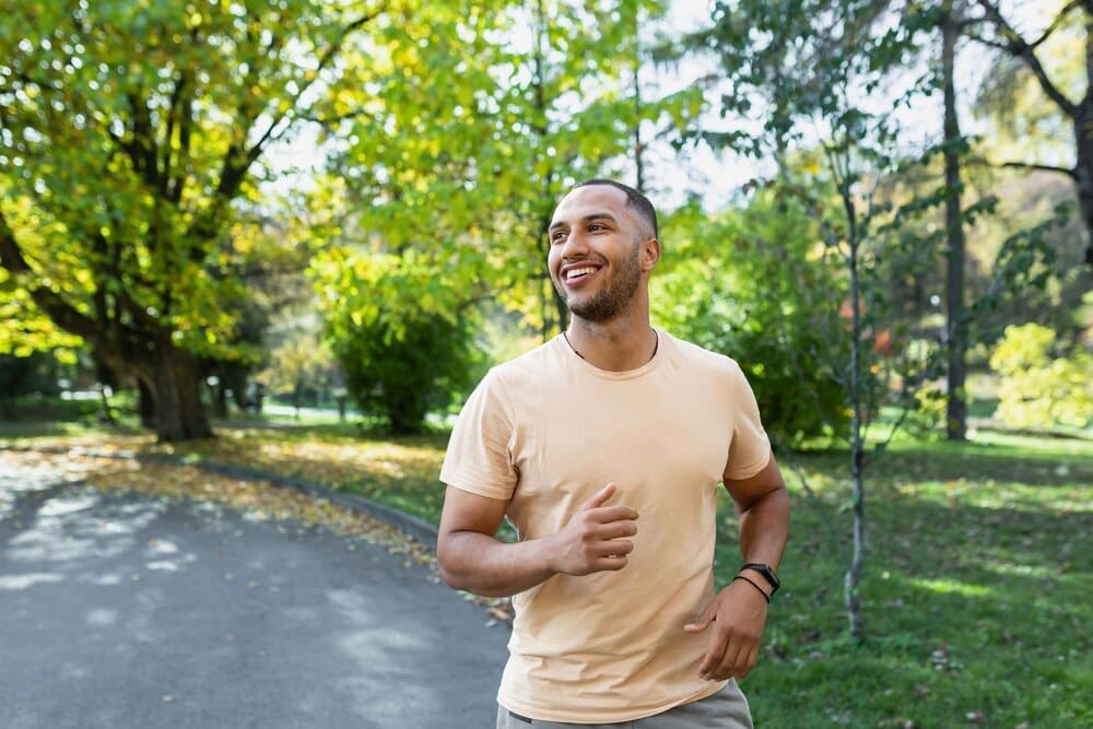 A smiling Hispanic man jogs through a sunny park.