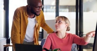 A Black man and woman in a corporate setting point at a computer screen while planning, possibly for a marketing project, with a mentor assisting the employee.