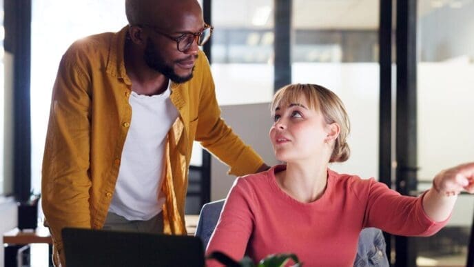 A Black man and woman in a corporate setting point at a computer screen while planning, possibly for a marketing project, with a mentor assisting the employee.