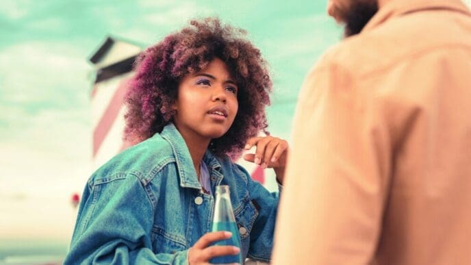 A young girl attentively listens to a young man while holding a glass bottle.