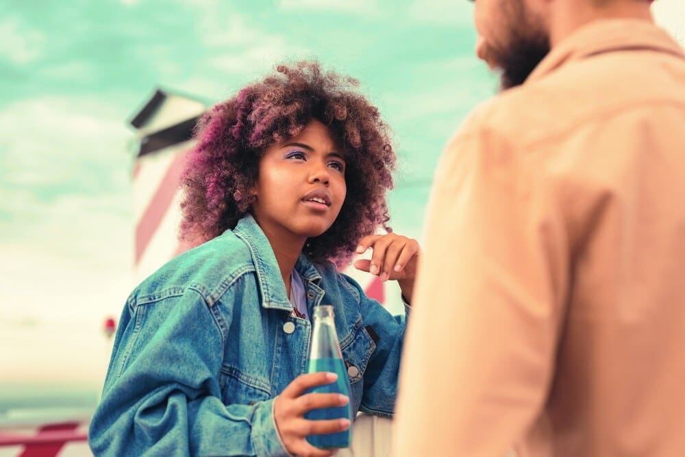 A young girl attentively listens to a young man while holding a glass bottle.
