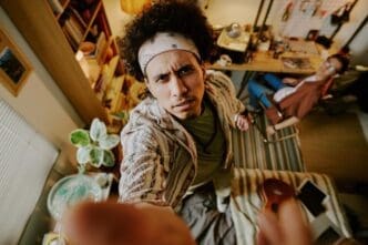 A man with curly hair and a black and white bandana installs a security camera while standing on a bed in a dorm room.