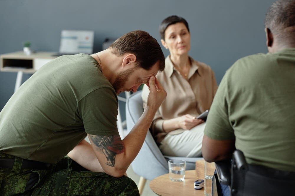 A soldier in uniform sits with their head in their hands, conveying a sense of sadness or despair.