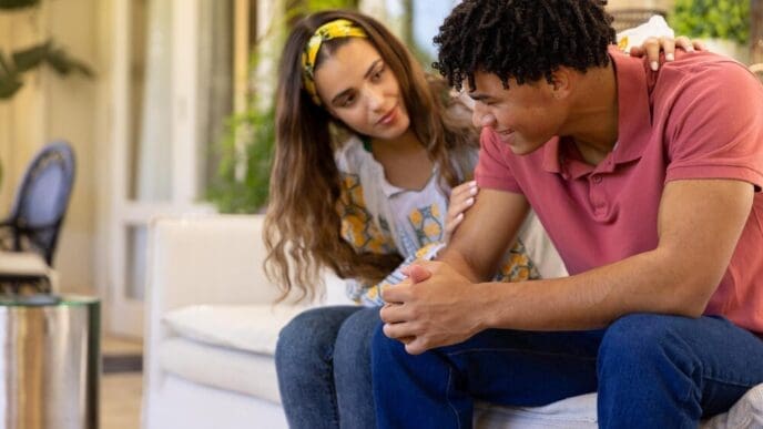 A diverse couple sits on a sofa in a living room, with the woman comforting the man.