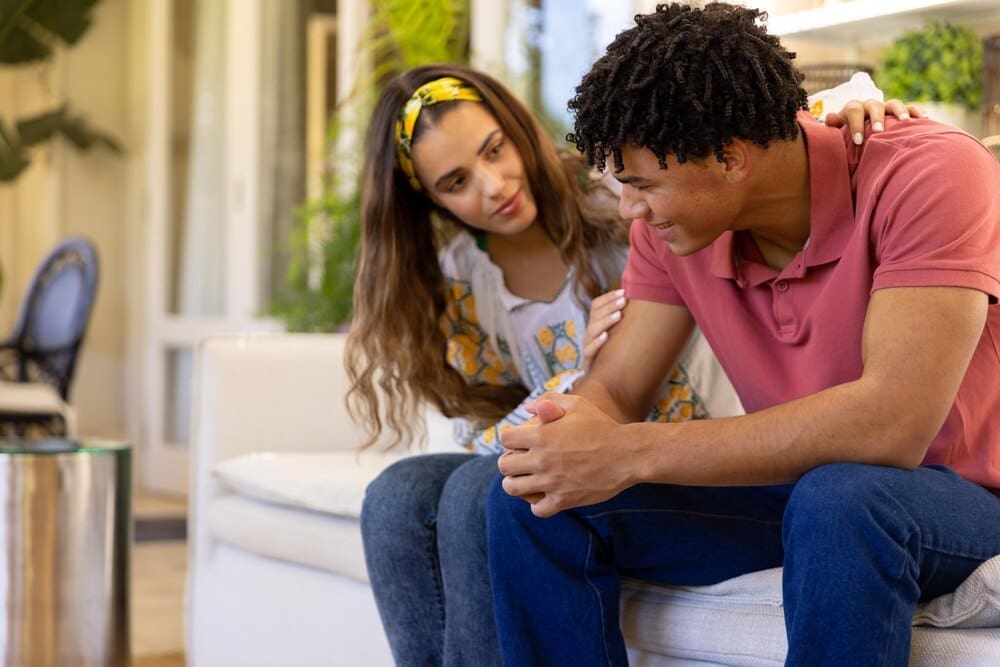 A diverse couple sits on a sofa in a living room, with the woman comforting the man.