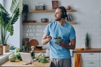 A young man in headphones leans on a kitchen counter, eating a fresh salad.