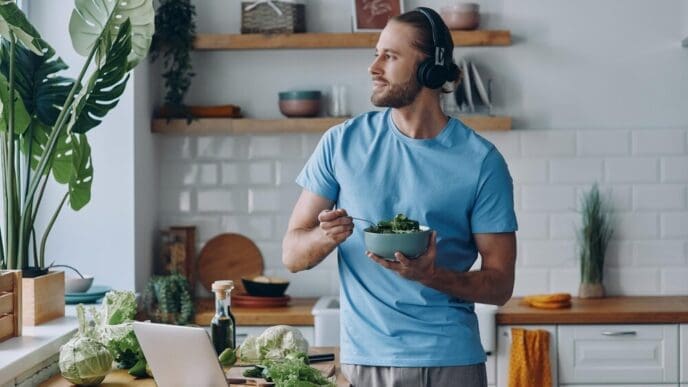 A young man in headphones leans on a kitchen counter, eating a fresh salad.