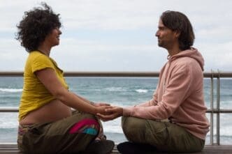 Expecting parents, seated on a bench in lotus position, hold hands on a beach in Las Palmas.
