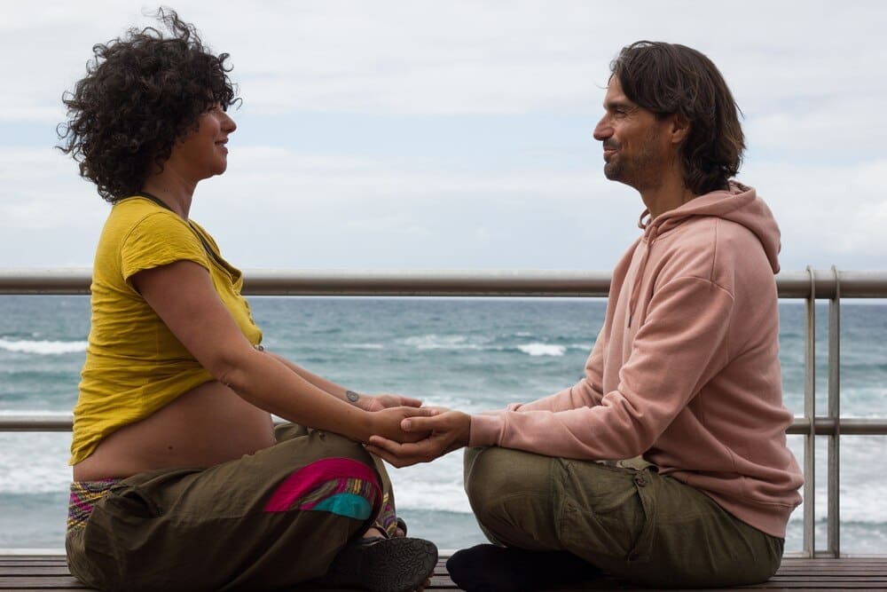 Expecting parents, seated on a bench in lotus position, hold hands on a beach in Las Palmas.