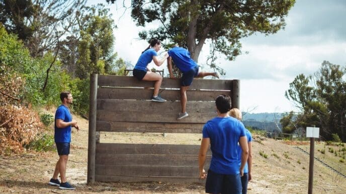 Female trainer helps a muscular man climb over a wooden wall on an outdoor obstacle course.
