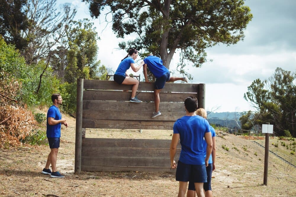 Female trainer helps a muscular man climb over a wooden wall on an outdoor obstacle course.