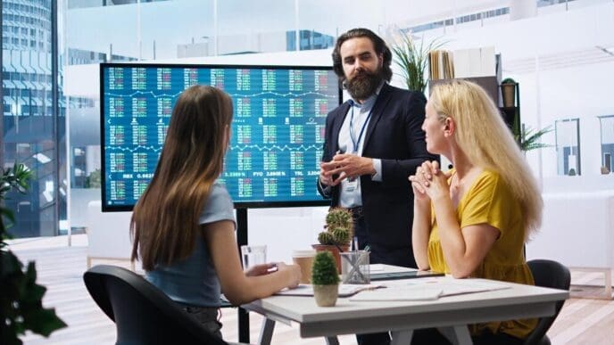 Financial advisor points at a stock market graph on a large screen while speaking to two clients.