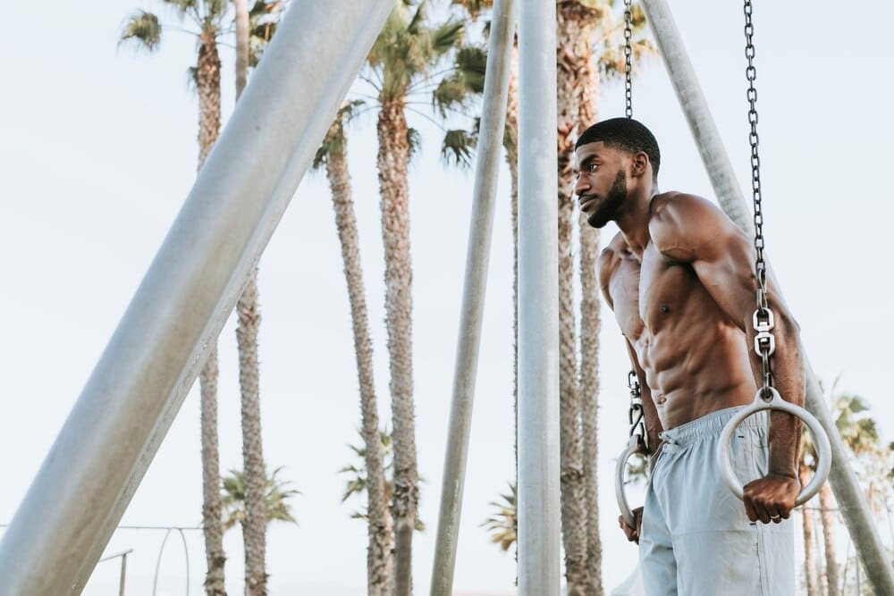 A muscular man does a workout on the beach, likely exercising with the ocean in the background.