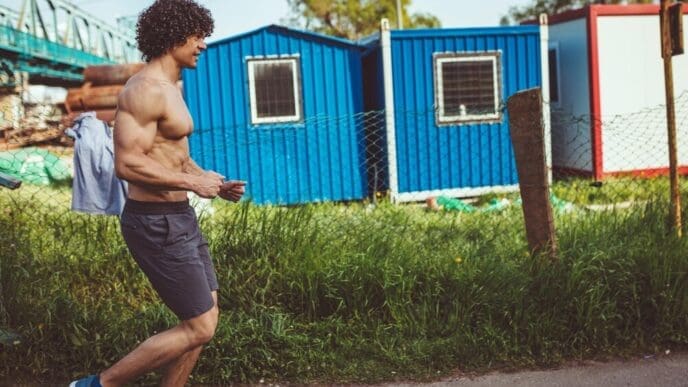 A shirtless young man with a muscular build runs through a city at dusk.