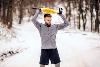 A sportsman stands on a snowy path in winter, lifting weights while exercising.