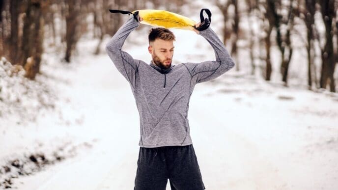 A sportsman stands on a snowy path in winter, lifting weights while exercising.
