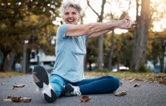 An elderly woman stretches on the ground in an outdoor fitness park.