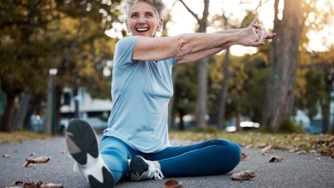 An elderly woman stretches on the ground in an outdoor fitness park.