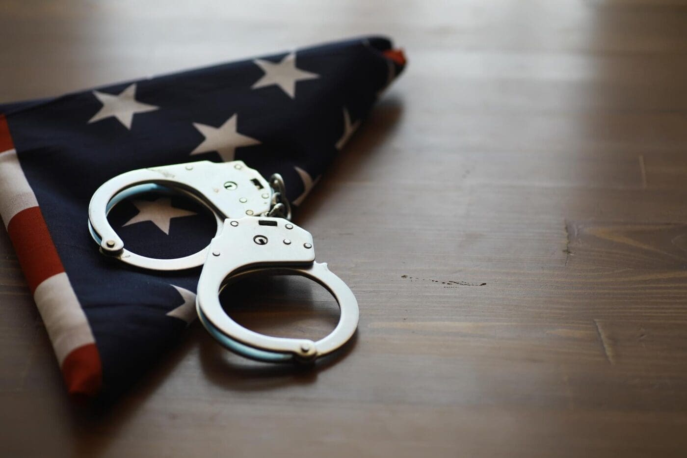 An American flag is folded and placed next to a pair of handcuffs on a wooden table, representing the intersection of law and patriotism.