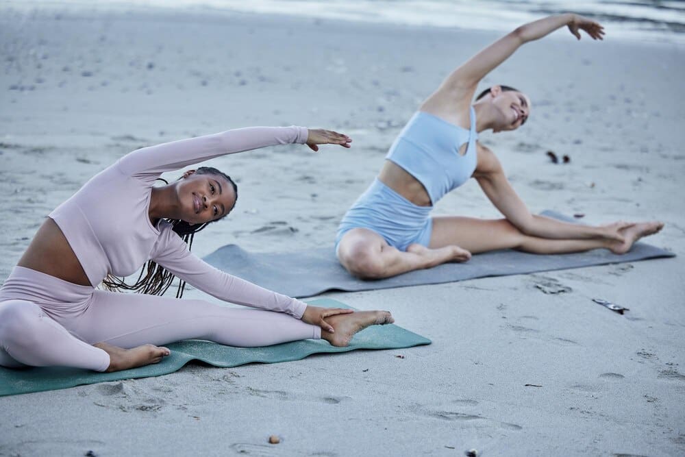 Two women smiling while practicing yoga and stretching on a beach.