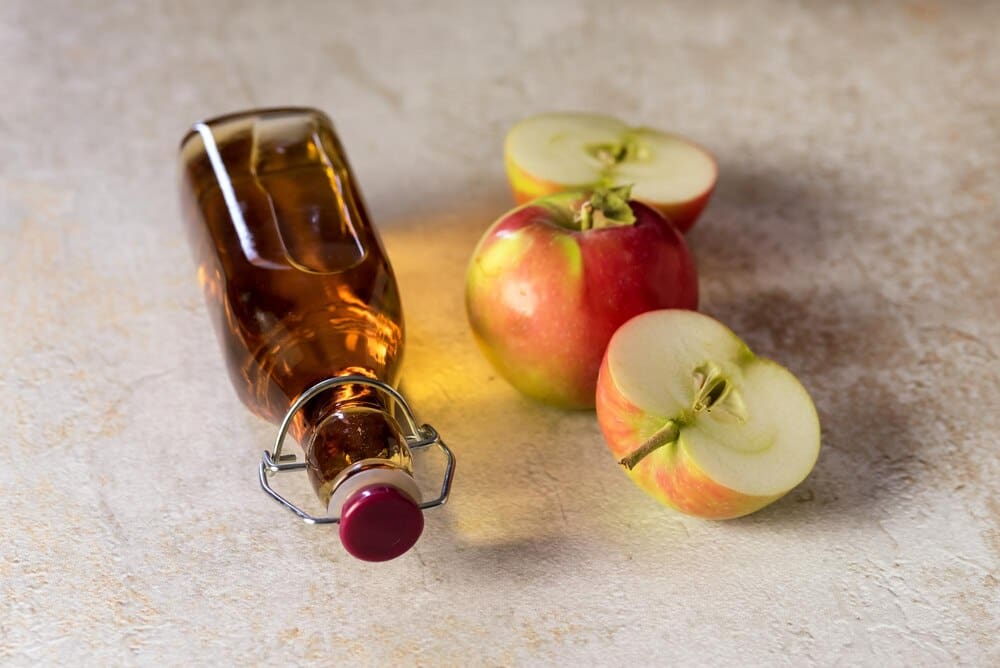 A glass bottle of apple vinegar and a fresh apple rest on a concrete surface, evoking a harvest theme.