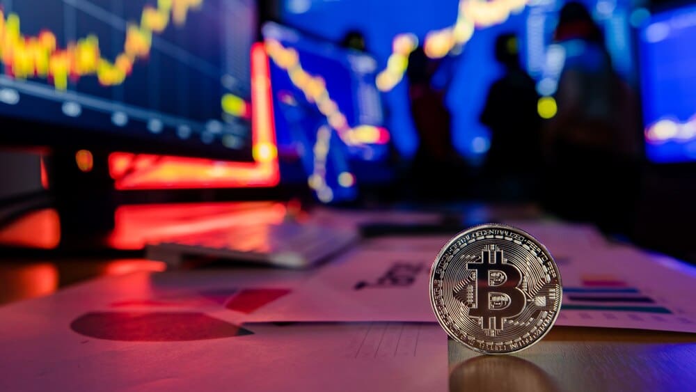 A golden Bitcoin token sits on a desk in front of a financial analysis graph charting trading activity.