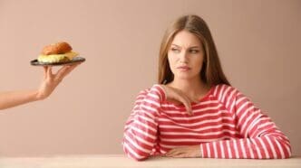 A person's hand offers a burger to a young woman seated at a table, who appears displeased.