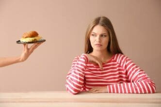 A person's hand offers a burger to a young woman seated at a table, who appears displeased.