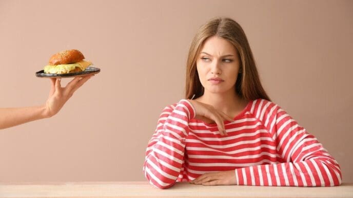 A person's hand offers a burger to a young woman seated at a table, who appears displeased.