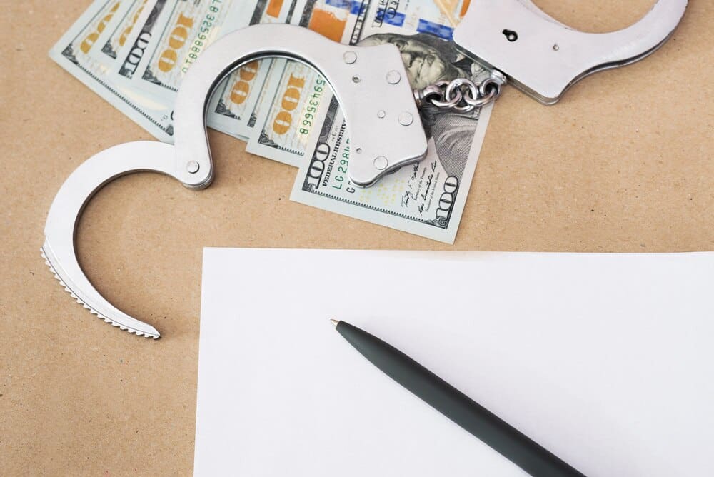 Handcuffs rest on a surface beside a stack of US dollar bills, a blank sheet of paper, and a pen.
