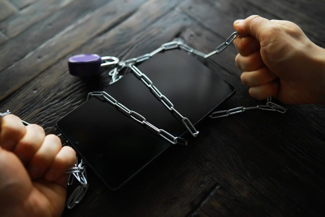 Hands bound with chains and a padlock encircling a smartphone, symbolizing digital addiction or security.