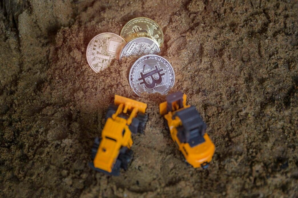 High-angle shot of a collection of Bitcoin cryptocurrency coins and toy trucks arranged on dirt.