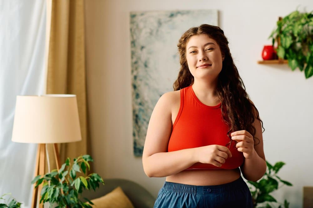 A smiling plus-size woman with curly hair wears comfortable clothes and enjoys the sunlight in her home.