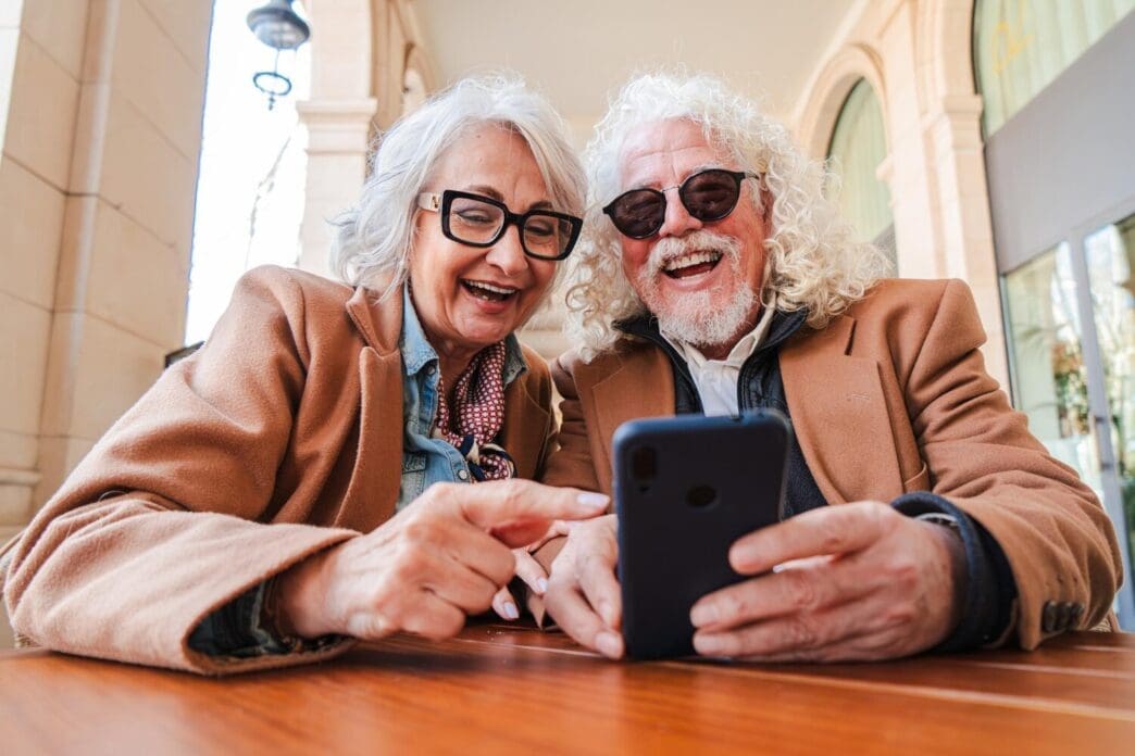 A smiling senior couple shares a smartphone, appearing joyful as they browse apps together.