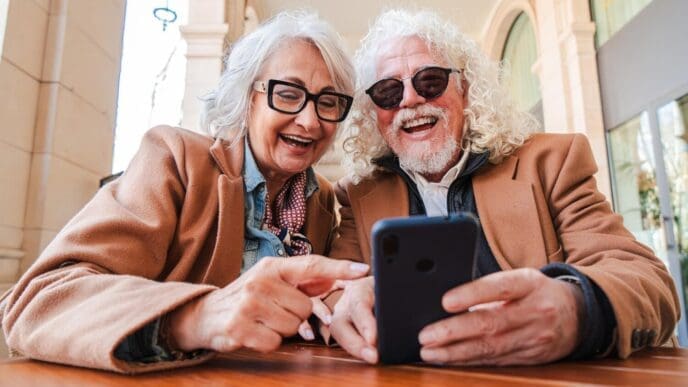 A smiling senior couple shares a smartphone, appearing joyful as they browse apps together.