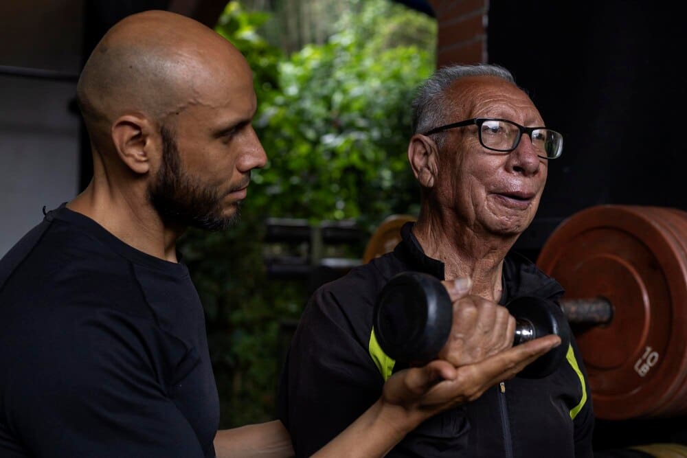 A senior Latin American man and his trainer do physical therapy exercises with a dumbbell in a gym.