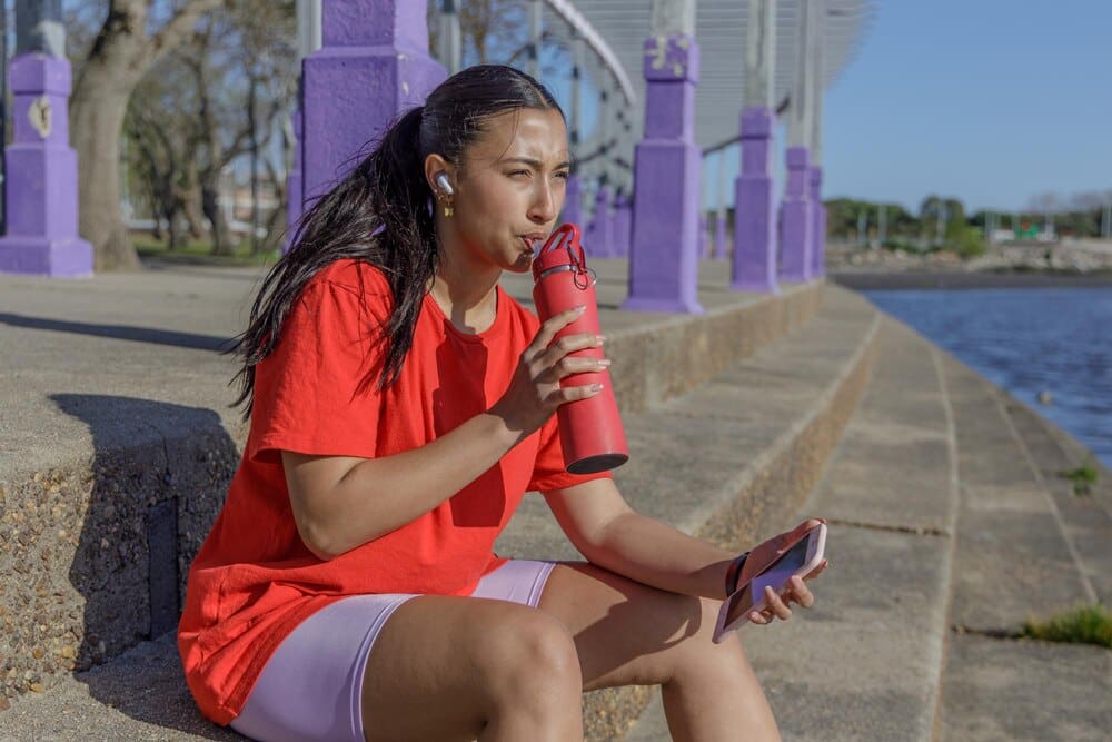 A Latina girl in sportswear sits on stairs, drinking from a water bottle.