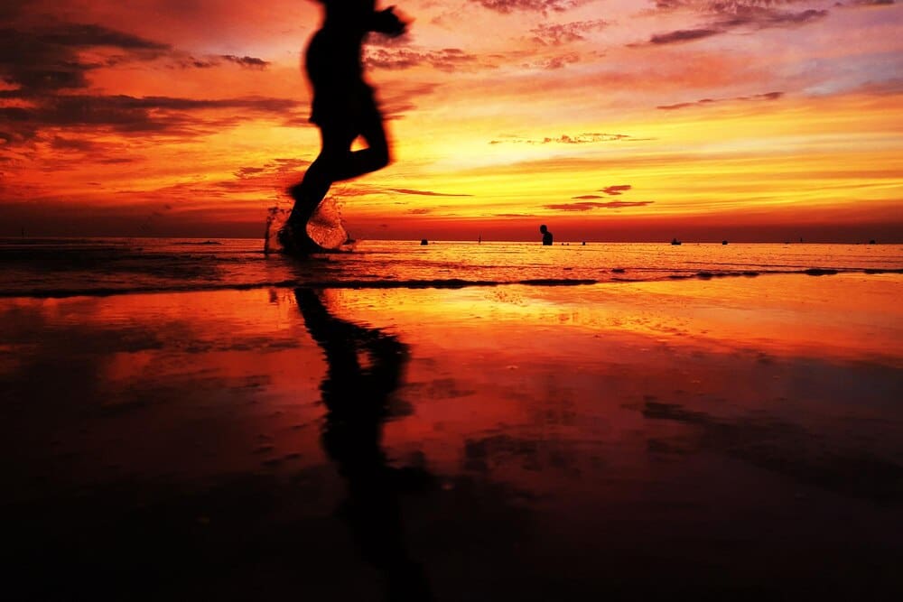 Low-angle view of a person running on a sandy beach, silhouetted against the vibrant colors of a sunset over the ocean.