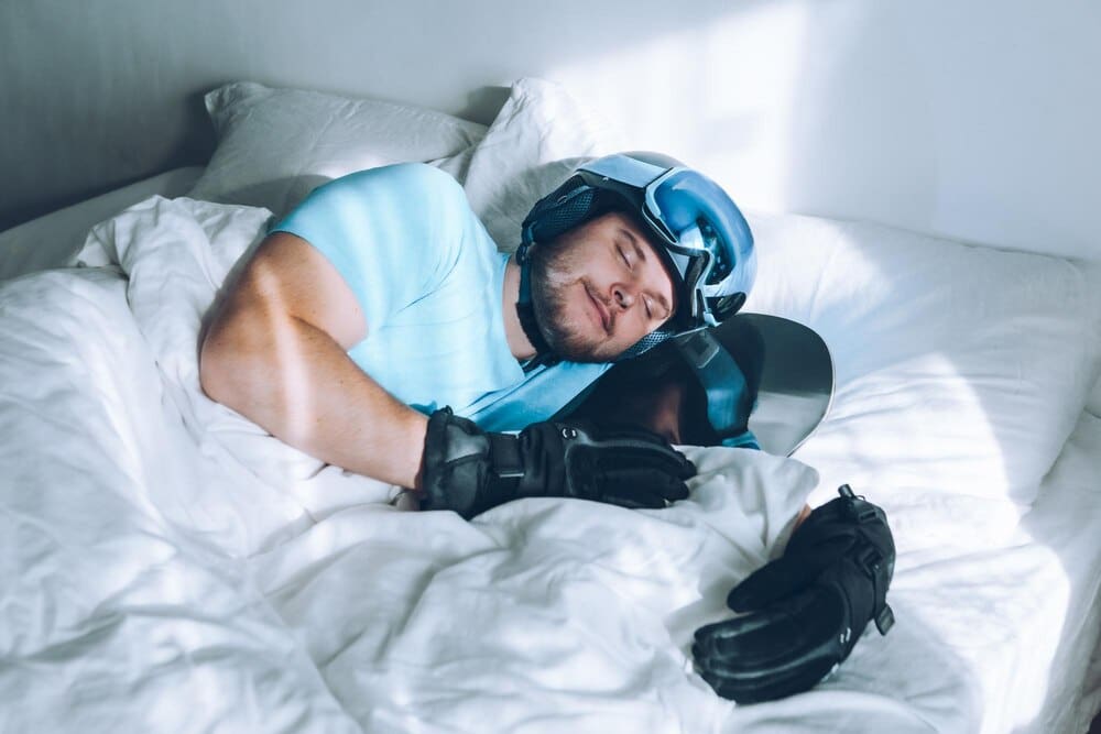 A man wearing ski goggles and a helmet lies in bed.