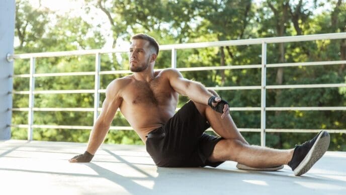 A muscular man with a naked torso stretches outdoors.