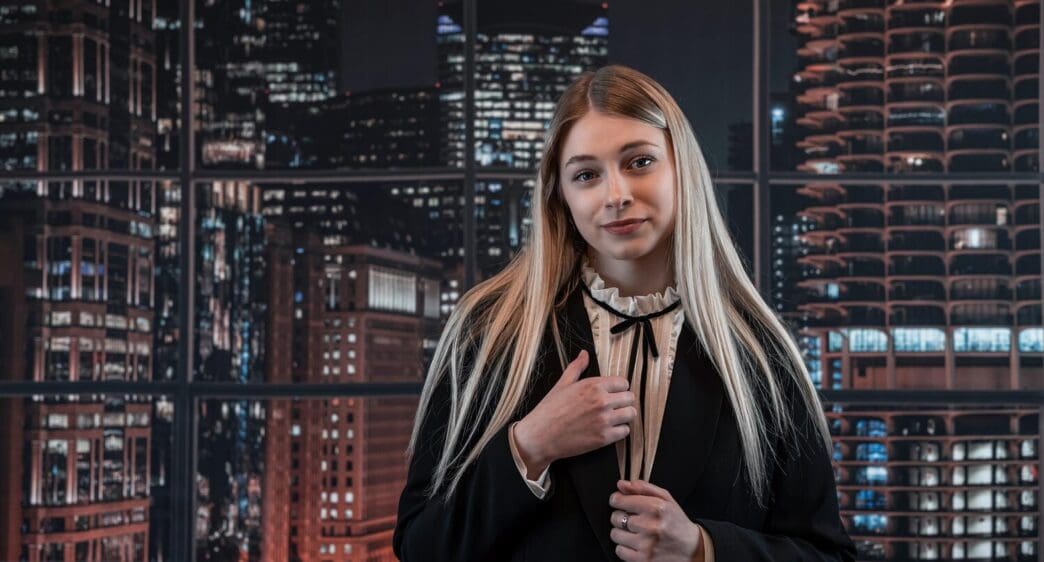 A young businesswoman in a black suit smiles confidently in an office setting.