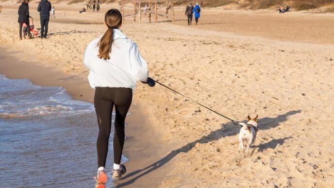 A girl runs on a beach with a dog, the sea in the background, and a few people walking in the distance, evoking a spring concept.