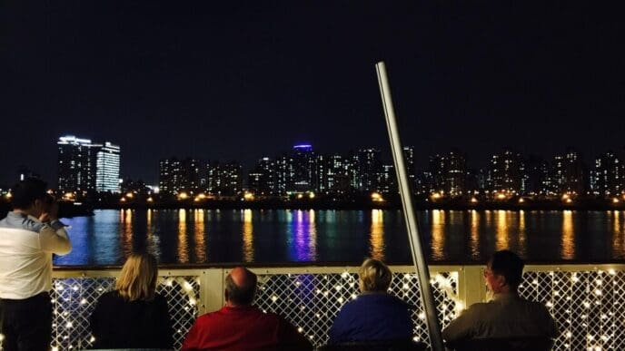 Rear view of people on a cruise ship looking at a brightly lit city skyline at night.