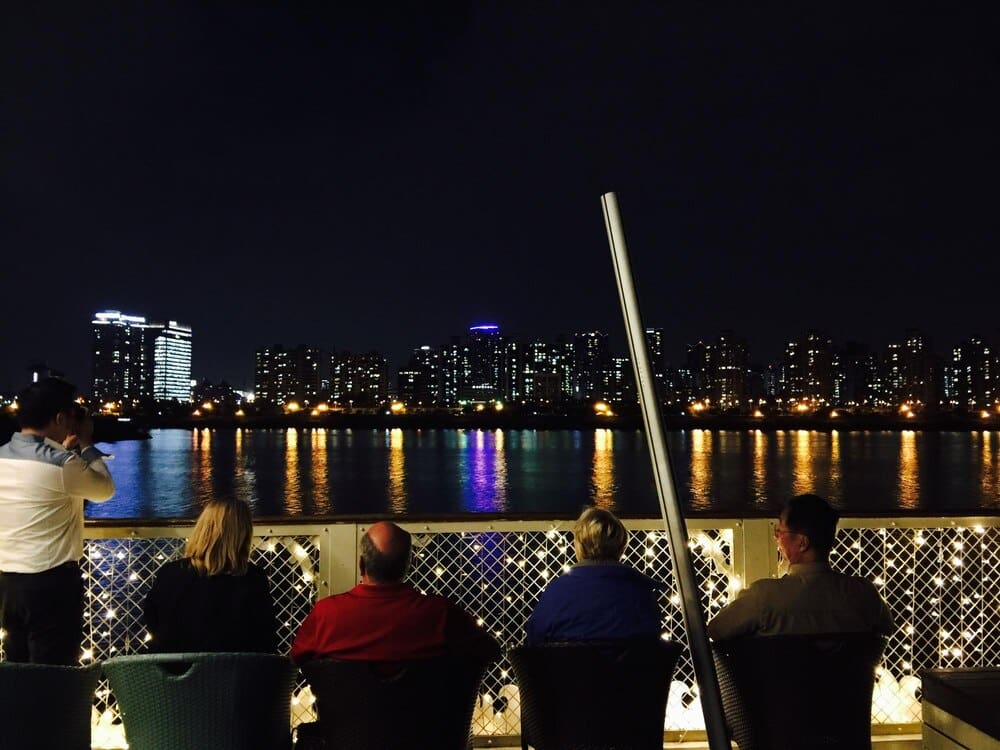 Rear view of people on a cruise ship looking at a brightly lit city skyline at night.