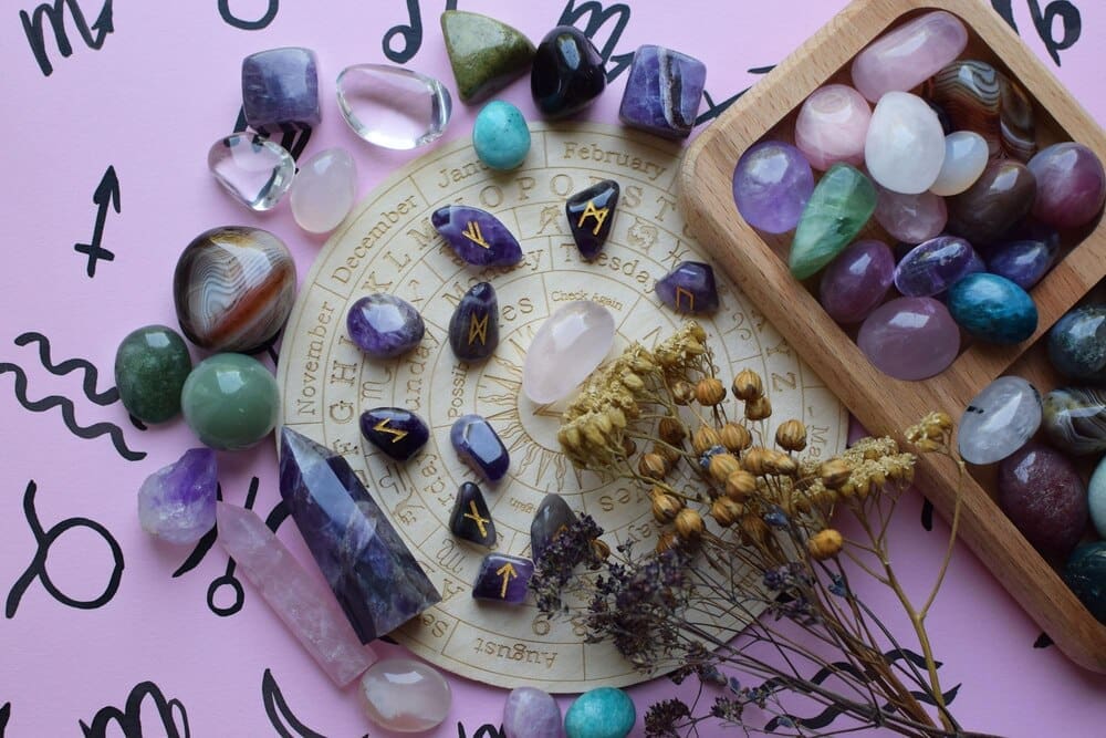 Amethyst runes and dried herbs are arranged on a pink surface.